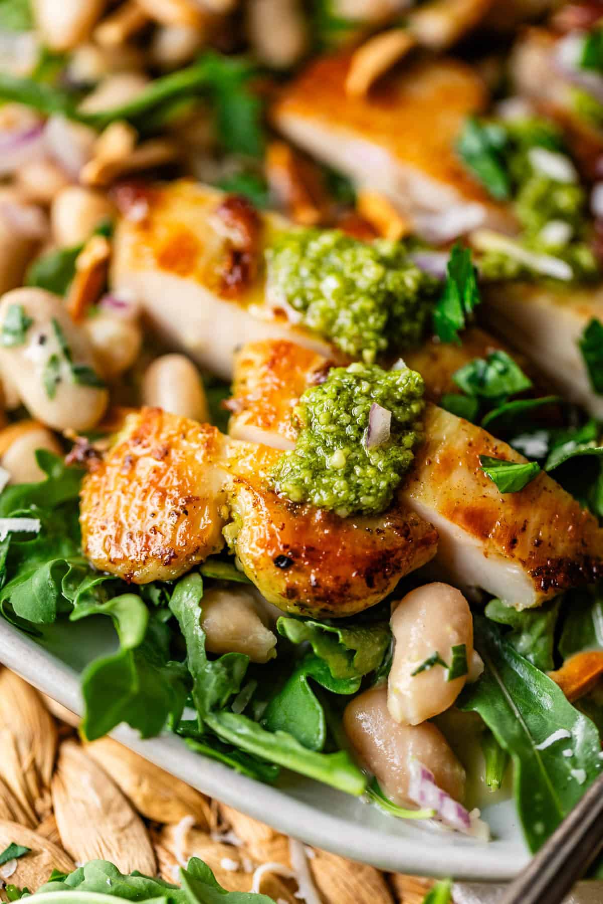 Close-up of sliced grilled chicken topped with green pesto, served on a bed of arugula, white beans, fresh herbs, and toasted almonds. The dish appears fresh and vibrant with a golden, crispy chicken skin.