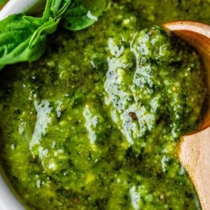 Close-up of vibrant green pesto sauce in a white bowl, garnished with fresh basil leaves. A wooden spoon rests in the sauce, showcasing its thick, textured consistency.