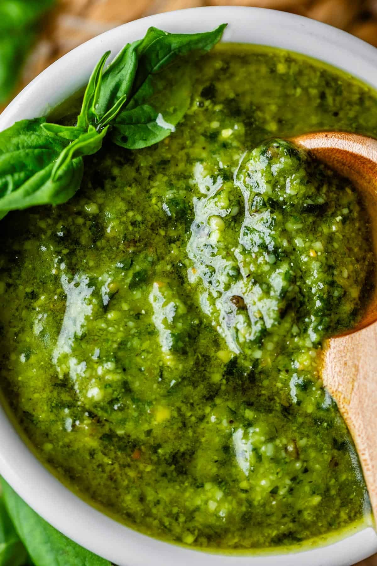 Close-up of vibrant green pesto sauce in a white bowl, garnished with fresh basil leaves. A wooden spoon rests in the sauce, showcasing its thick, textured consistency.