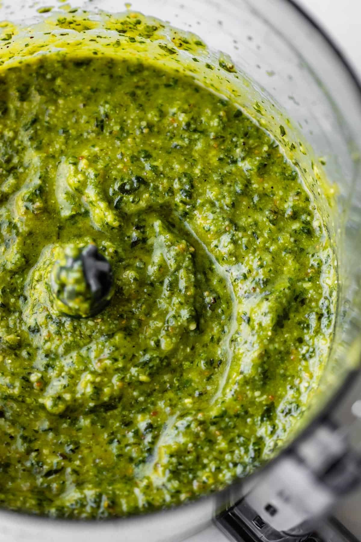 A close-up view of vibrant green pesto sauce being blended in a food processor, showing a creamy, textured consistency with visible flecks of herbs and nuts.
