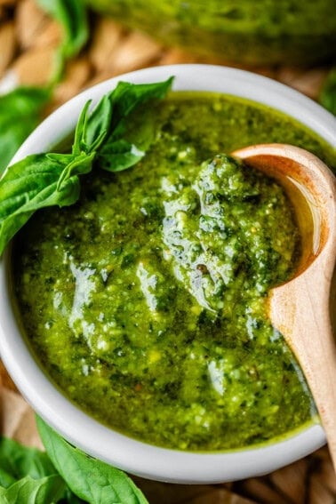 A white bowl filled with green pesto sauce, garnished with fresh basil leaves, with a wooden spoon resting inside. The pesto appears chunky and glossy.