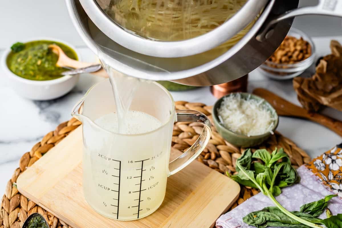 A pot of hot pasta water is being poured into a large measuring jug on a woven mat, surrounded by fresh basil, grated cheese, pesto in a bowl, and scattered kitchen utensils.