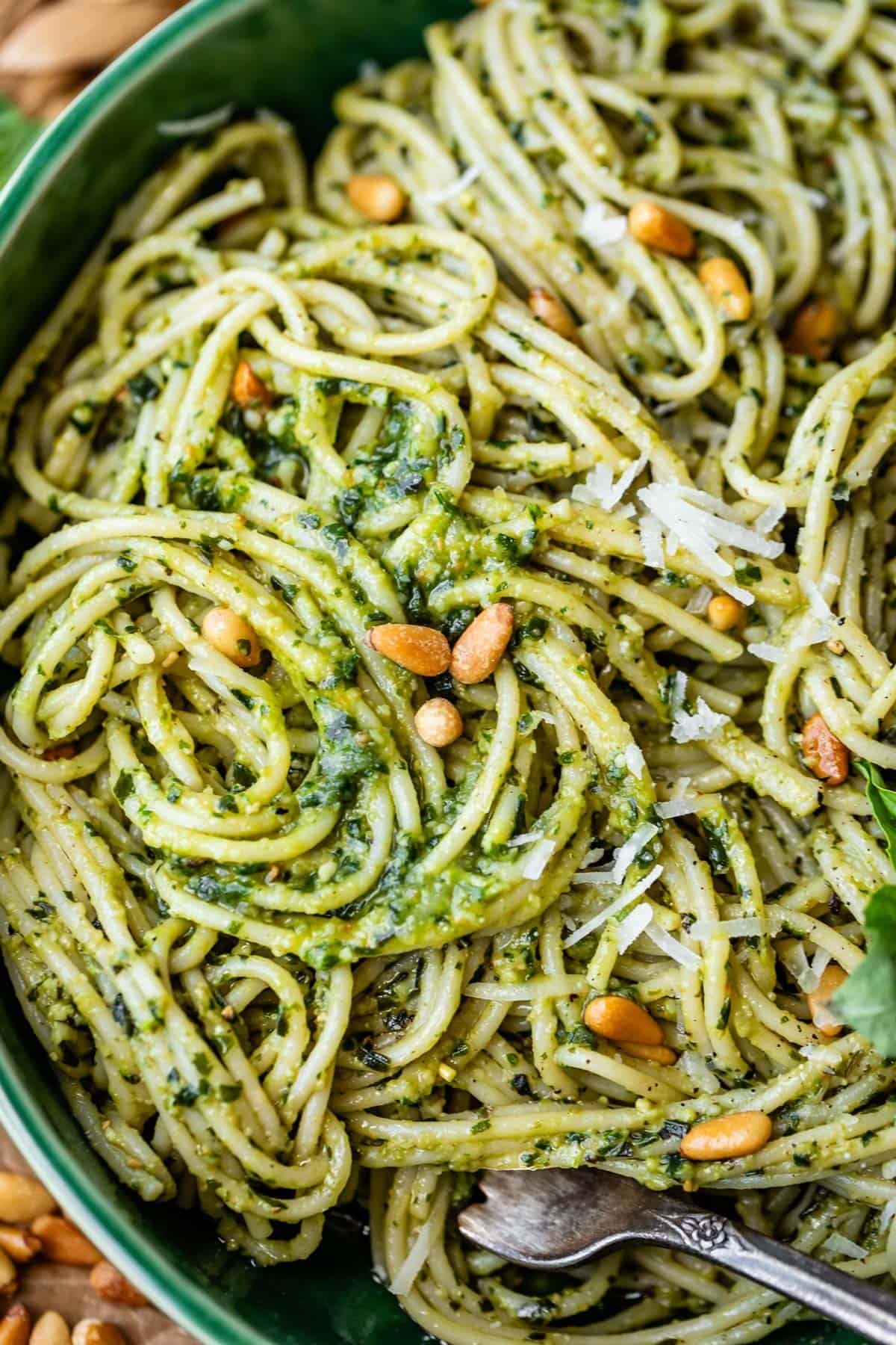 Close-up of spaghetti tossed in green pesto sauce, garnished with pine nuts and grated Parmesan cheese, served in a bowl with a fork.