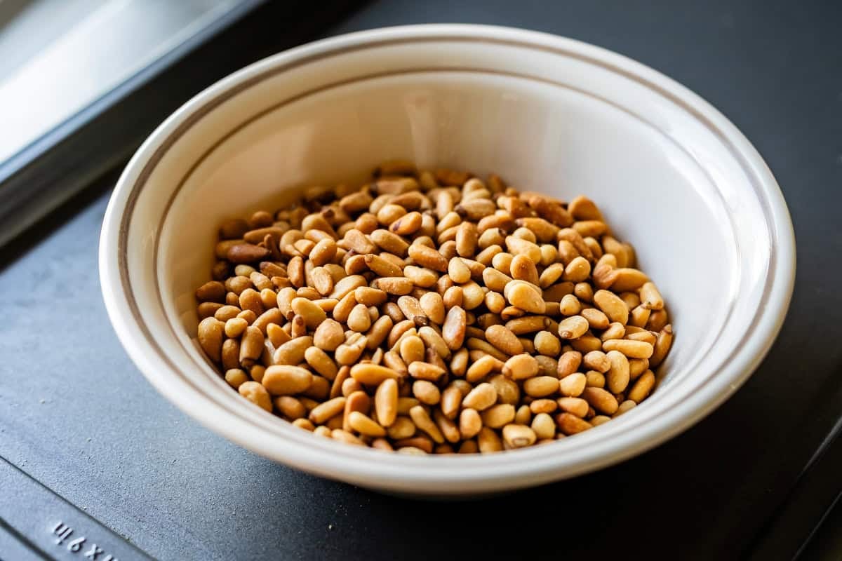 A white bowl filled with toasted pine nuts sits on a dark surface.