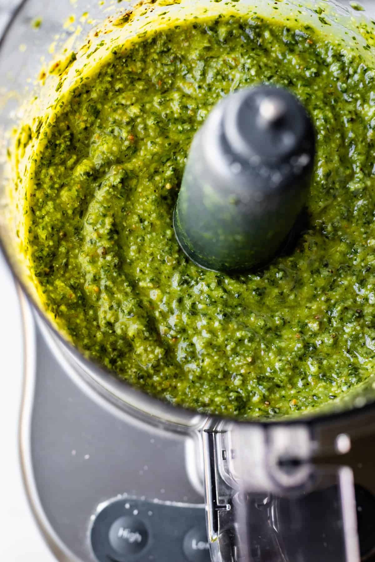 A close-up of vibrant green pesto being blended in a food processor, with the mixture swirling around the central blade and the texture visibly creamy and slightly chunky.