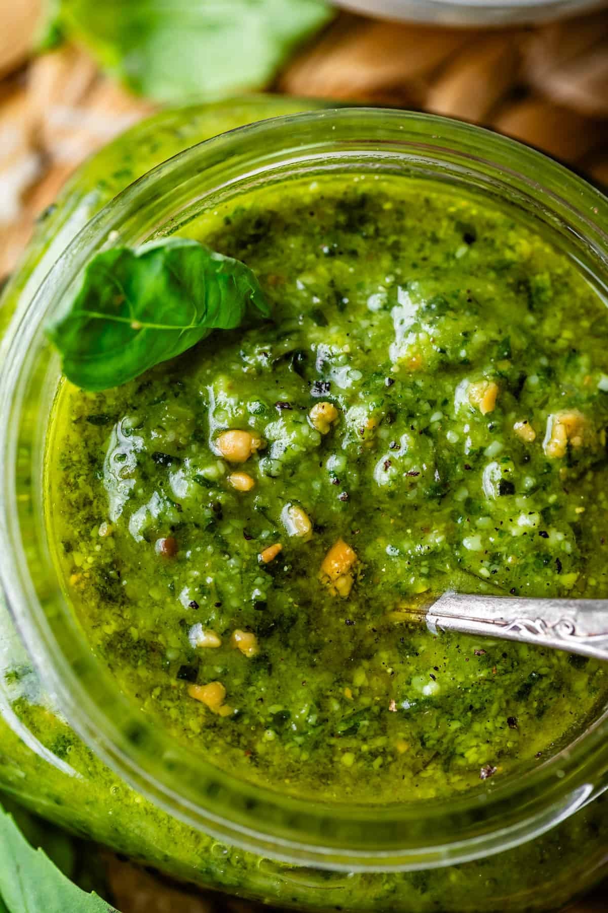 Close-up of a jar filled with vibrant green pesto sauce, garnished with a fresh basil leaf. A spoon is partially dipped in the textured sauce, showing bits of nuts and herbs throughout.