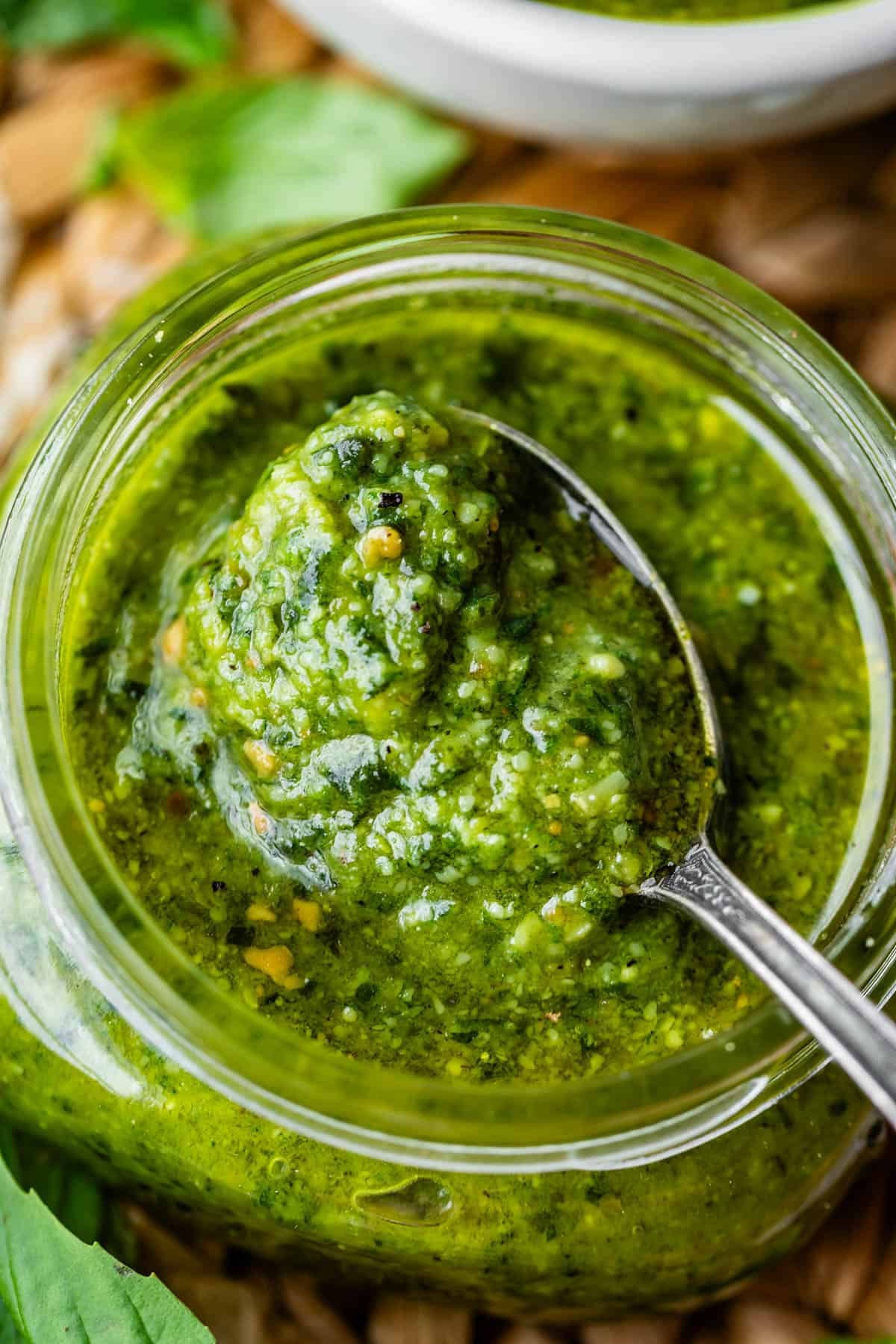 Close-up of a jar of vibrant green pesto sauce with a spoonful being lifted from the jar; fresh herbs are visible in the background.