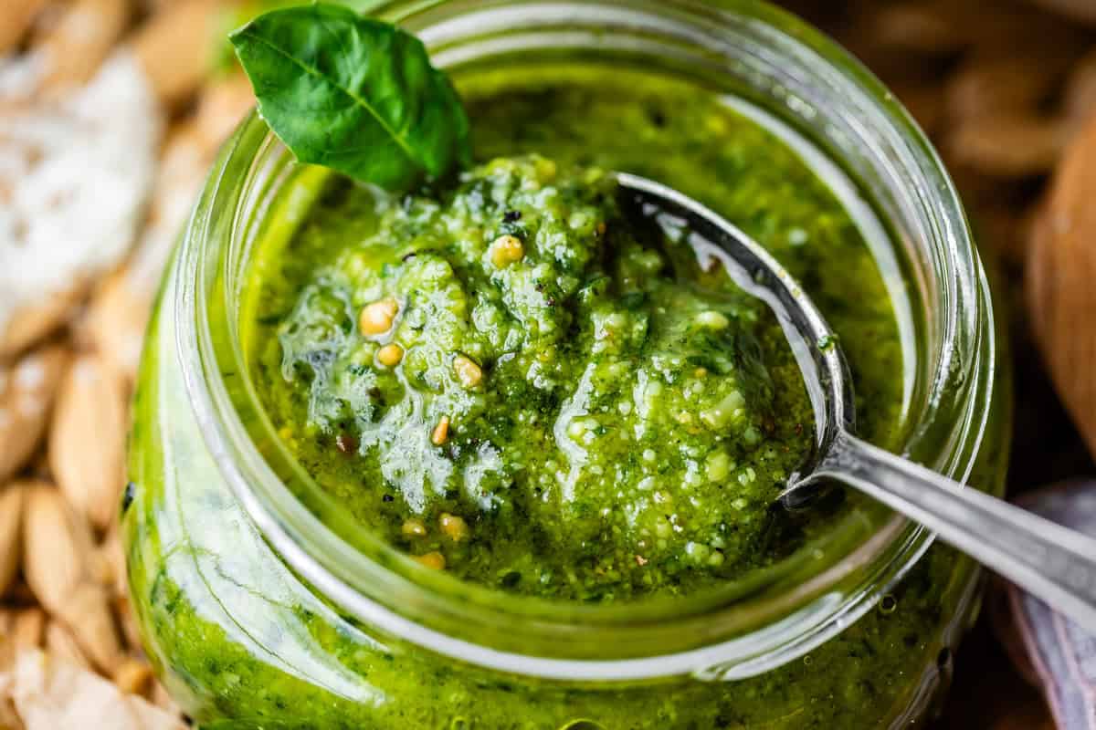 A close-up of a glass jar filled with green pesto sauce. A spoon is inside the jar, and a fresh basil leaf garnishes the top. The pesto appears chunky, with visible seeds and herbs.