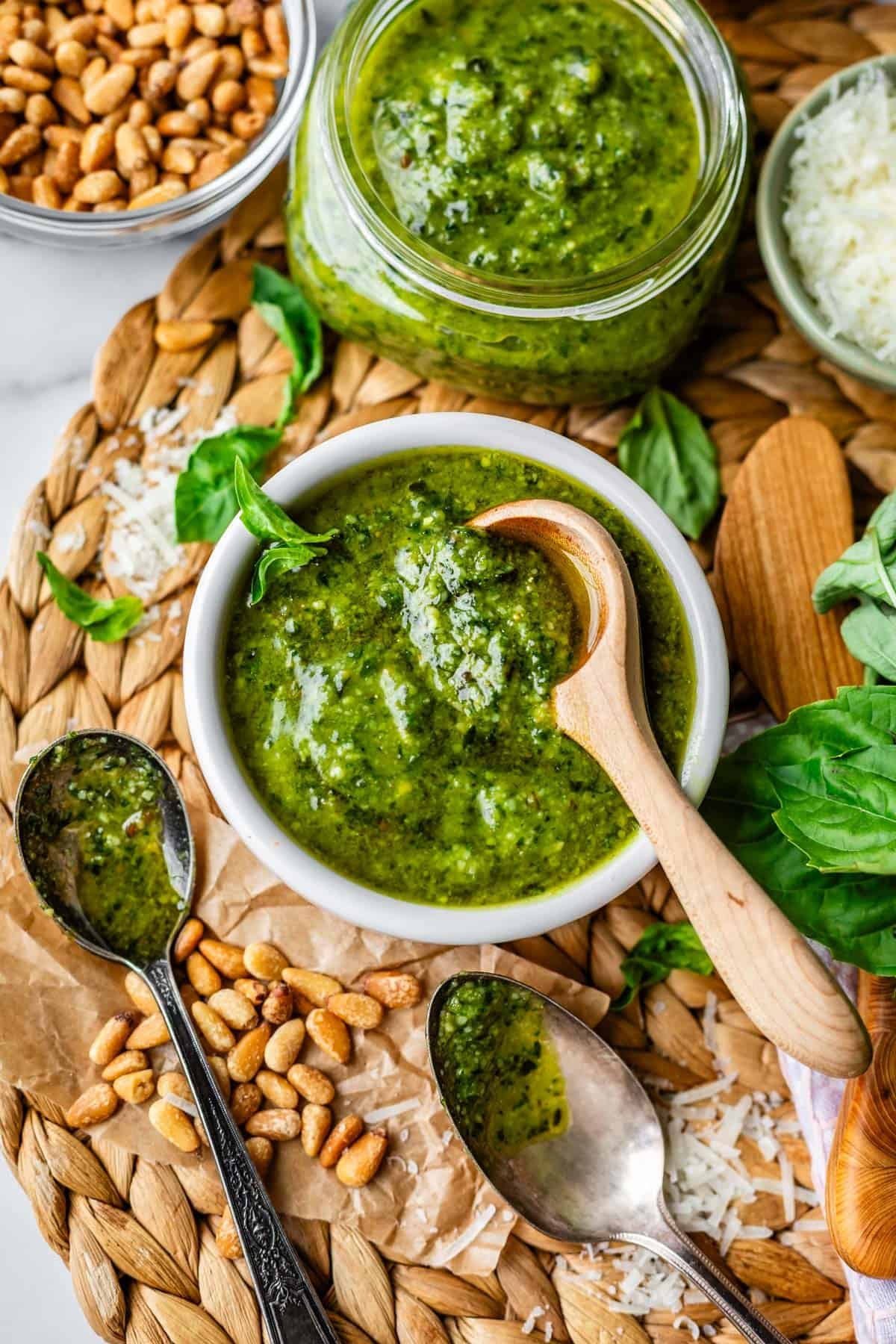 A bowl of green pesto with a wooden spoon, surrounded by pine nuts, fresh basil leaves, grated cheese, and a glass jar of pesto on a woven placemat.