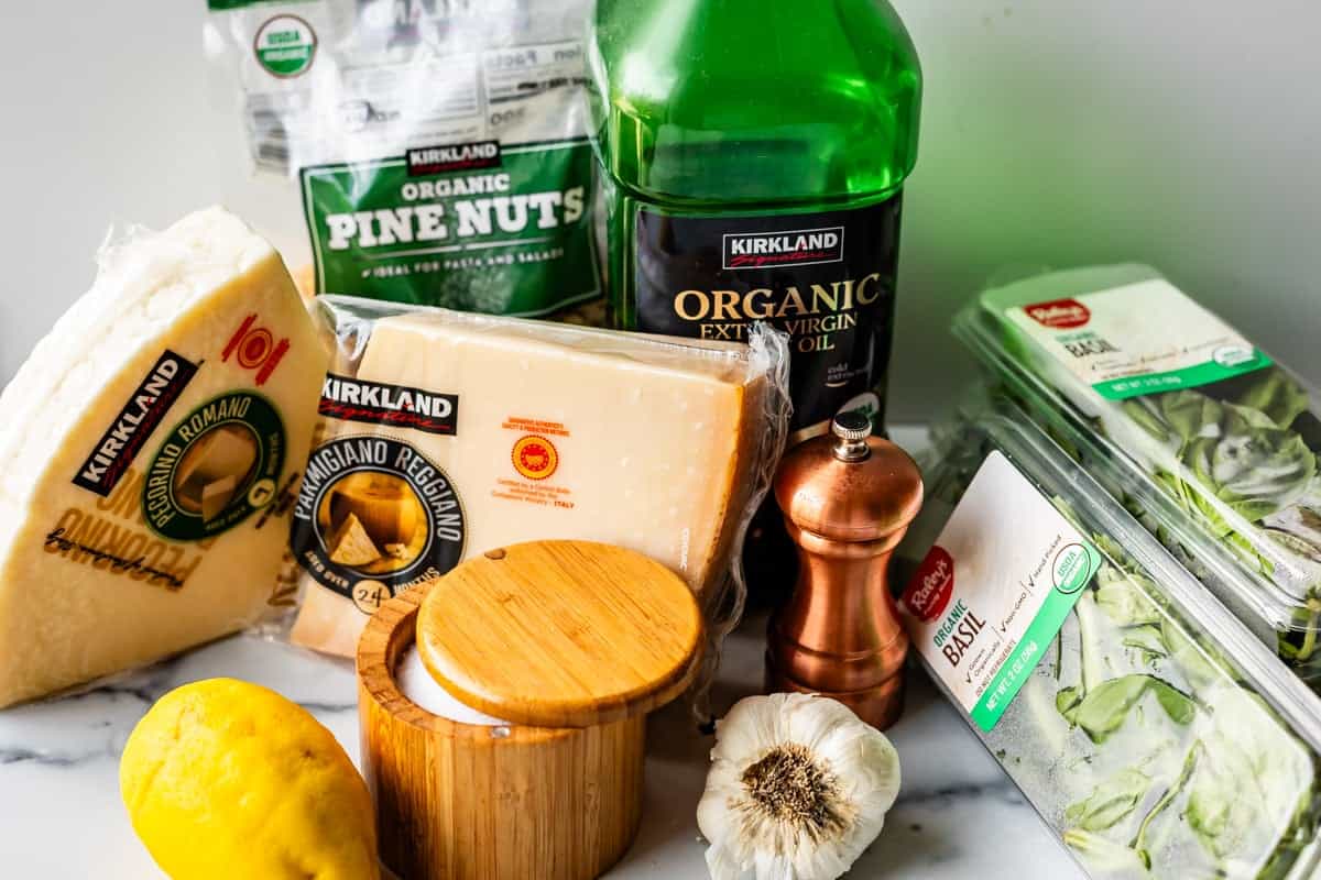 A collection of ingredients for pesto, including parmesan and romano cheese wedges, organic pine nuts, extra virgin olive oil, fresh basil, a lemon, garlic bulb, salt cellar, and a wooden pepper grinder on a marble surface.