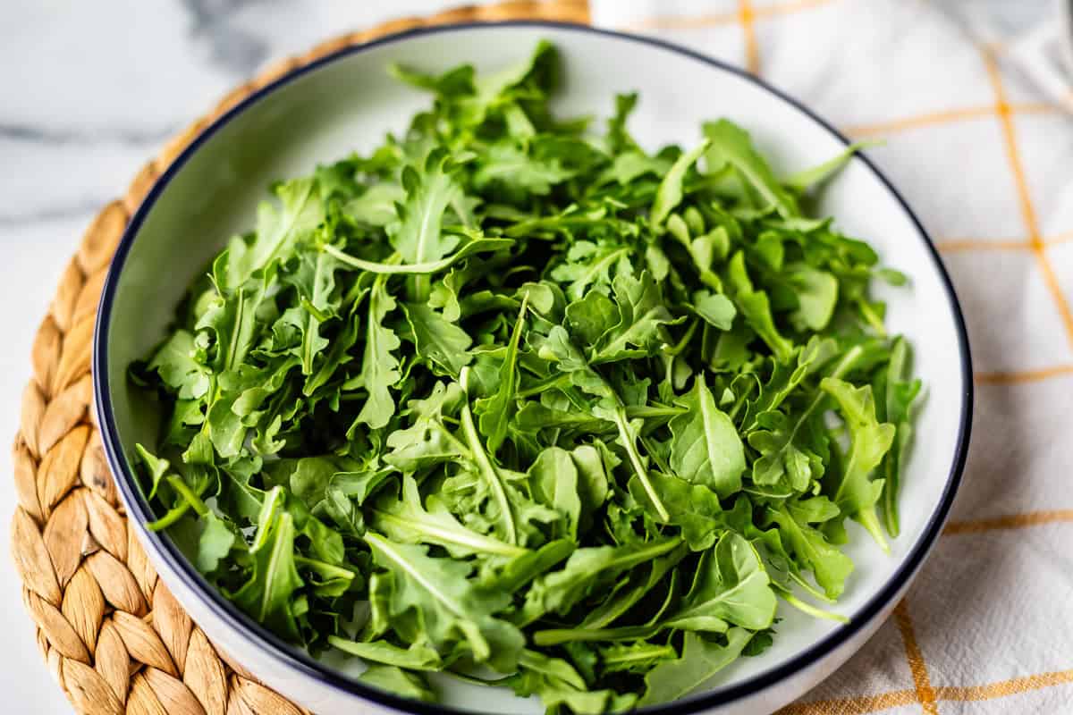 A white bowl filled with fresh green arugula leaves sits on a woven placemat with part of a white cloth visible nearby.