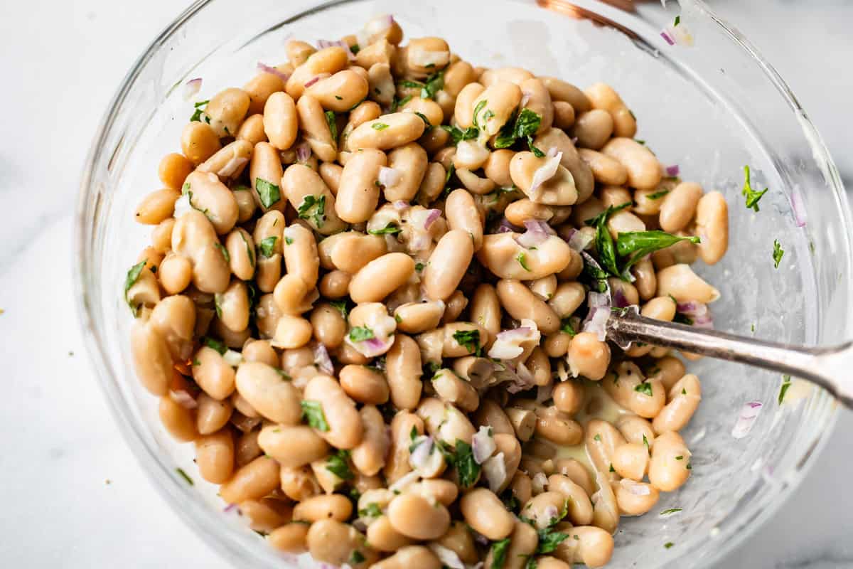 A clear glass bowl filled with a bean salad made of white beans, chopped red onions, and fresh parsley, with a metal spoon resting inside.