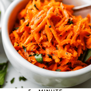 A white bowl filled with shredded carrot salad, garnished with fresh herbs and black pepper, with a spoon inside. A small bowl of chopped herbs and another bowl of salad are blurred in the background. Text reads: 5-Minute French Carrot Salad.
