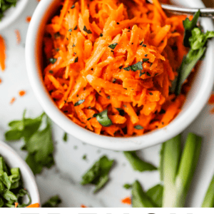 A bowl of shredded French carrot salad garnished with herbs, surrounded by small dishes of chopped greens and utensils on a white surface. Text at the bottom reads “French Carrot Salad The Food Charlatan.”.