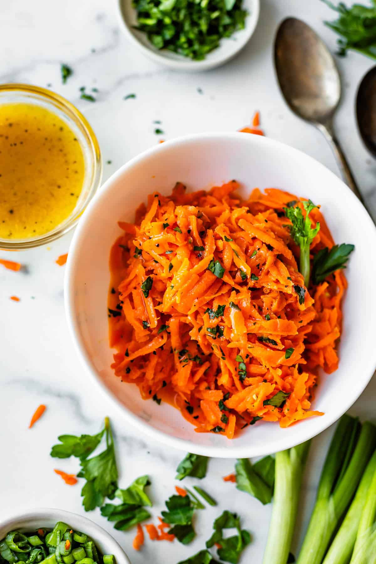 A white bowl filled with shredded carrot salad garnished with parsley, surrounded by a small bowl of dressing, a spoon, and chopped herbs on a white surface.