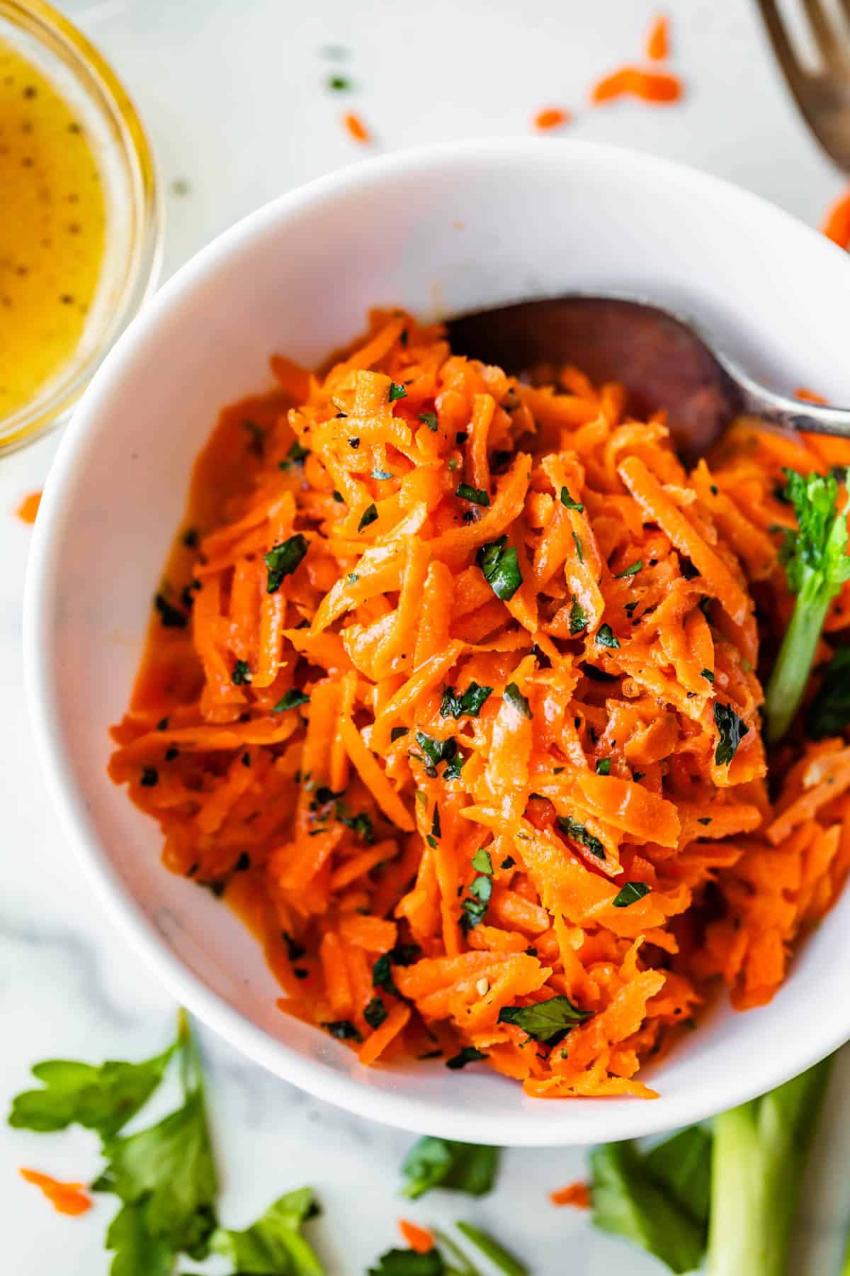 A white bowl filled with shredded carrot salad garnished with chopped herbs, with a spoon inside the bowl. Fresh parsley and a small glass container of dressing are nearby on the table.