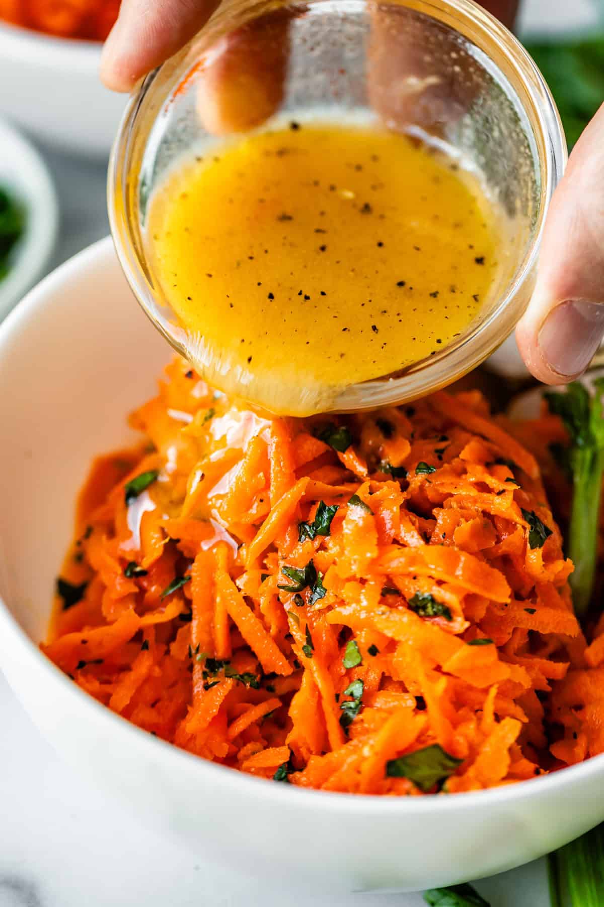 A hand pours a small bowl of vinaigrette dressing over a bowl of shredded carrots mixed with fresh herbs.