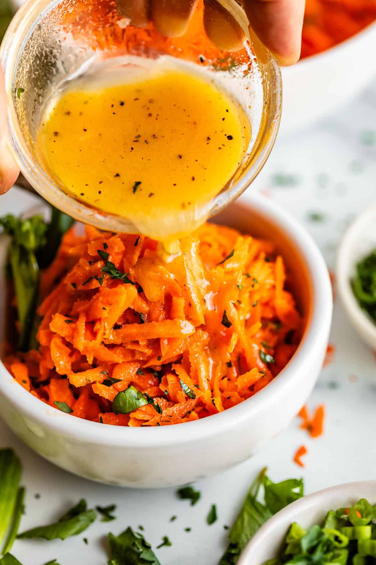 A hand pours a small bowl of vinaigrette dressing over a serving of shredded carrot salad garnished with chopped herbs, in a white bowl on a white surface with herbs scattered around.