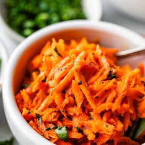 A white bowl filled with shredded carrot salad garnished with chopped herbs and freshly ground black pepper, with a spoon in the bowl and blurred bowls of greens in the background.