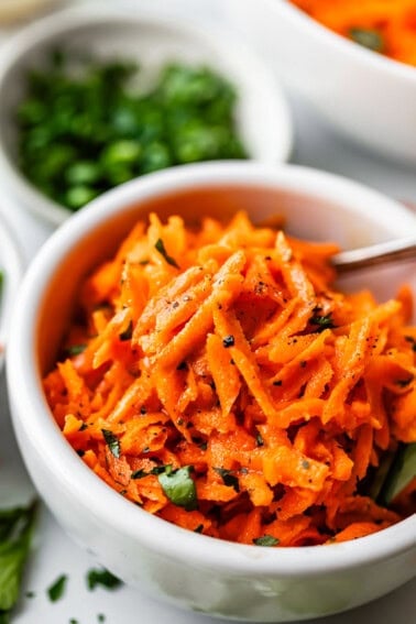 A white bowl filled with shredded carrot salad garnished with chopped herbs and freshly ground black pepper, with a spoon in the bowl and blurred bowls of greens in the background.
