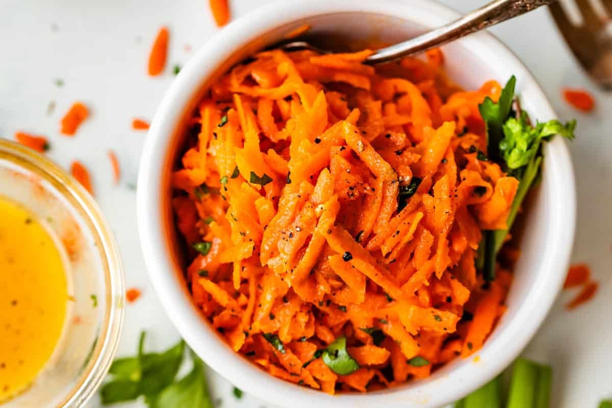 A white bowl filled with shredded carrot salad garnished with herbs and cracked black pepper, with a spoon in the bowl. Scattered carrot shreds and a small bowl of yellow dressing are nearby.
