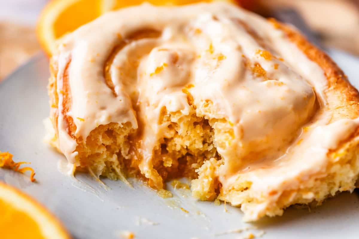 Close-up of a partially eaten orange cinnamon roll topped with creamy icing, served on a light-colored plate with orange slices in the background.