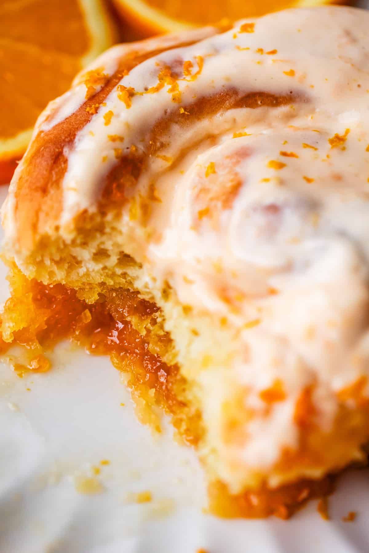 A close-up of a partially eaten orange roll topped with creamy orange glaze and orange zest, with bright orange slices blurred in the background.