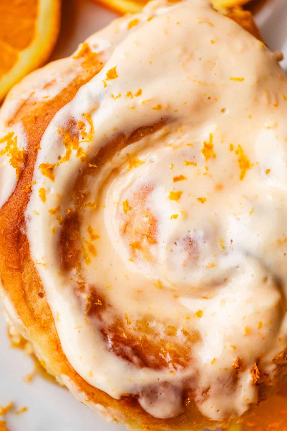 A close-up of an orange roll topped with creamy orange icing and sprinkled with orange zest, with a slice of orange visible in the background.