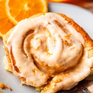 A close-up of a cinnamon roll topped with creamy orange glaze, sitting on a white plate with fresh orange slices in the background.
