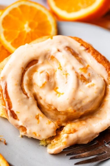 A close-up of a cinnamon roll topped with creamy orange glaze, sitting on a white plate with fresh orange slices in the background.