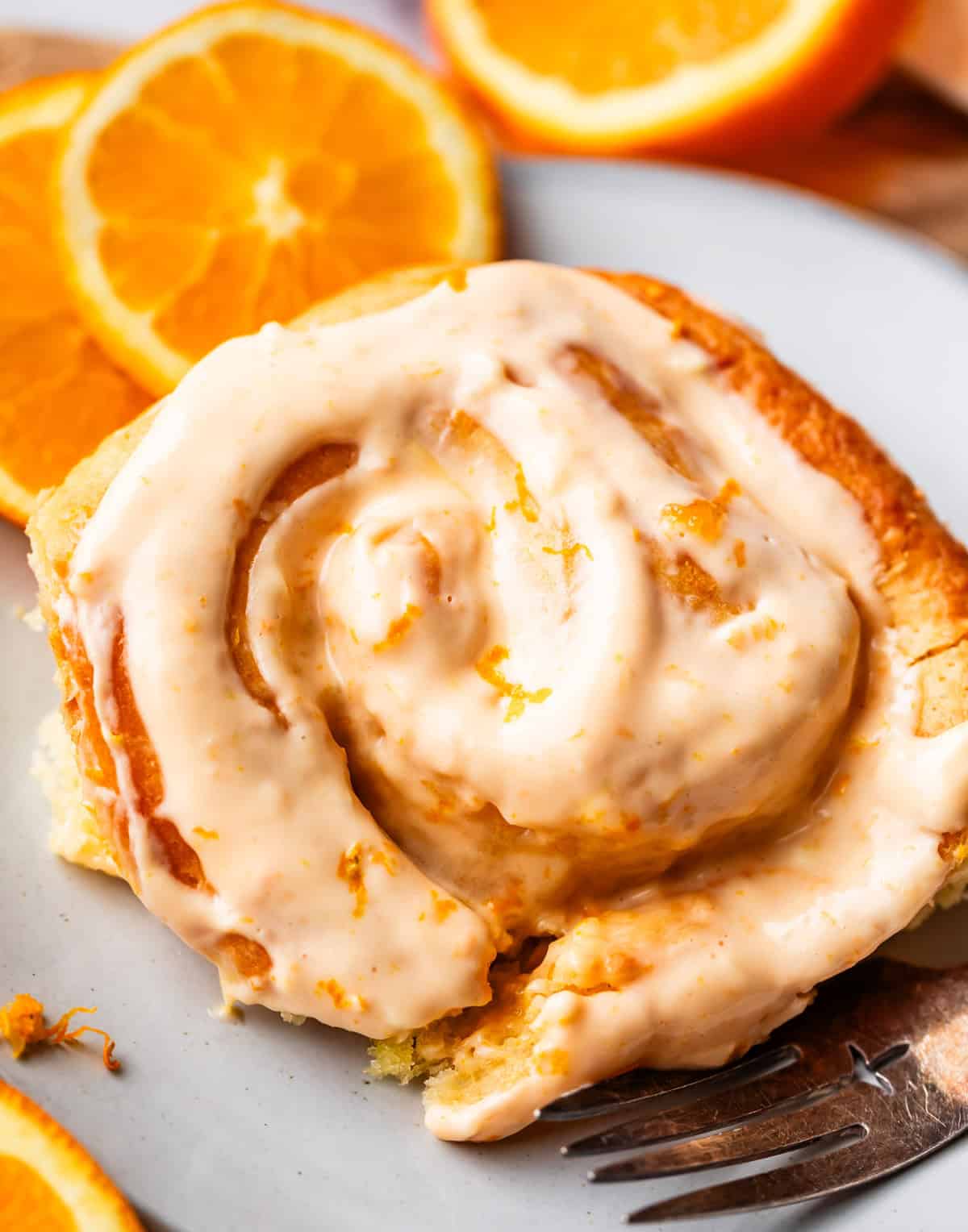 A close-up of a cinnamon roll topped with creamy orange glaze, sitting on a white plate with fresh orange slices in the background.