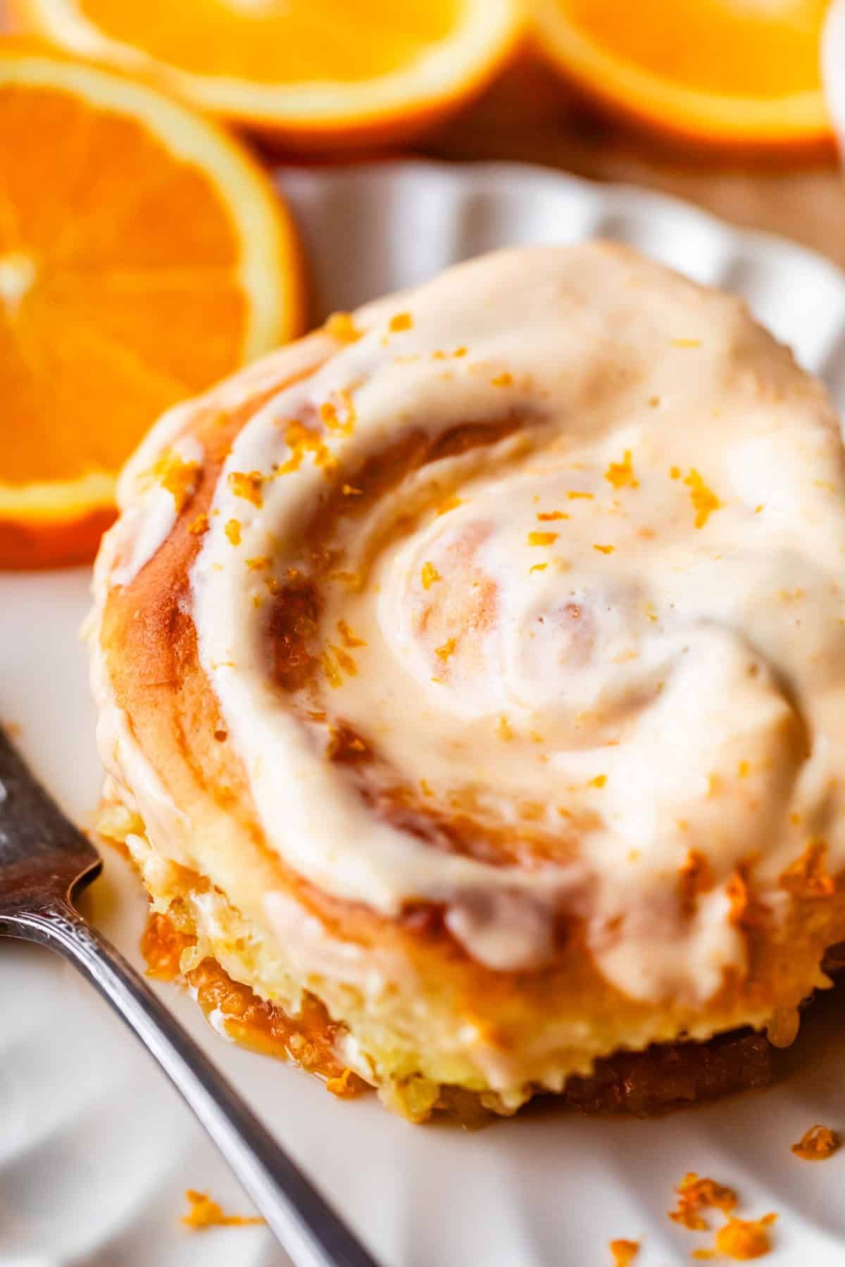 A close-up of an orange sweet roll topped with creamy icing and orange zest, served on a white plate with a fork next to it and fresh orange slices in the background.