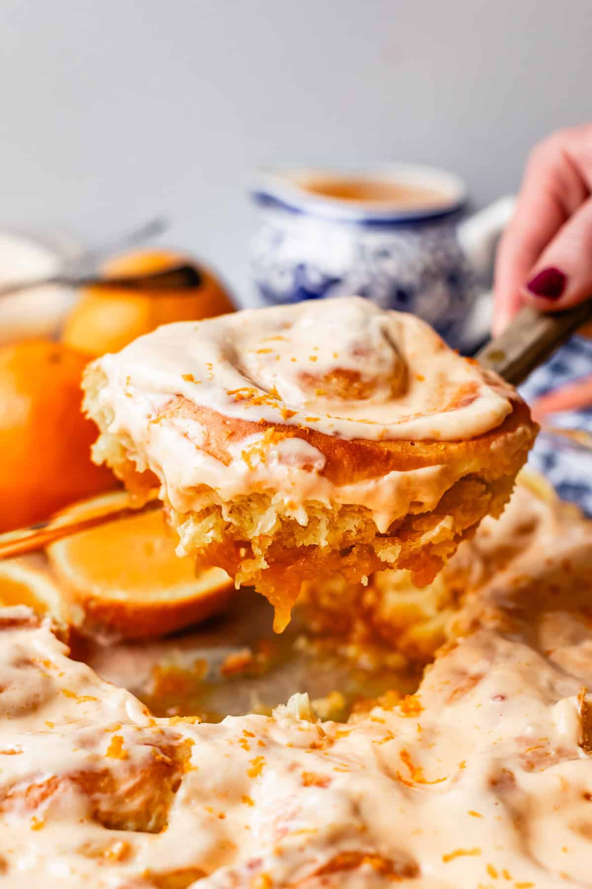 A hand lifts a frosted cinnamon roll topped with orange zest from a tray of similar rolls. In the background, there are oranges, a cup, and a blurred blue-and-white teapot.