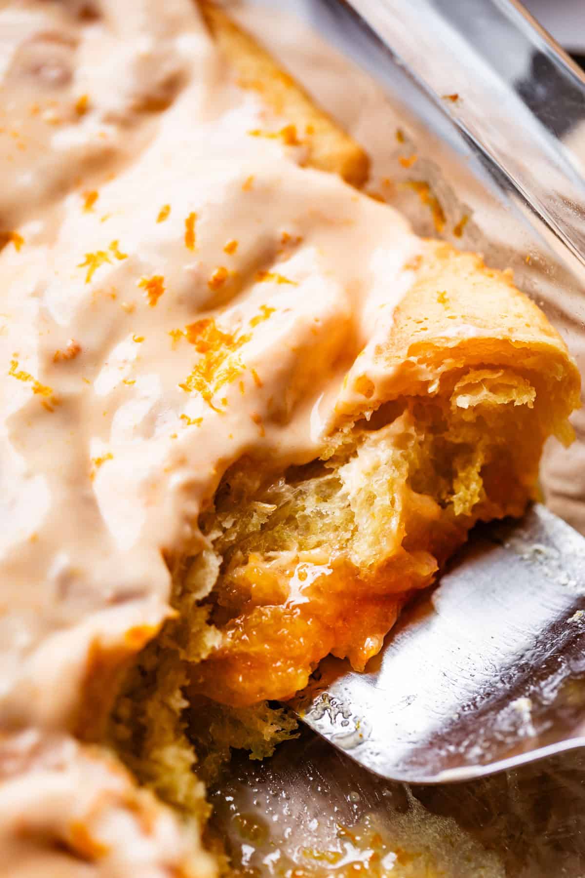 A close-up of a glazed pastry being lifted with a metal spatula, revealing a gooey orange filling and topped with cream-colored icing and orange zest.
