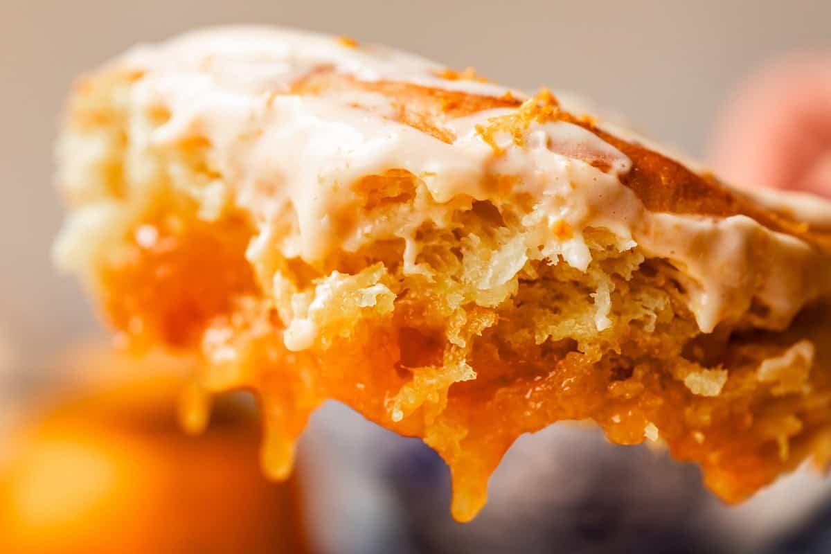Close-up of a flaky orange roll with a gooey orange filling oozing out, topped with a creamy orange glaze. The background is softly blurred, focusing attention on the texture and layers of the dessert.