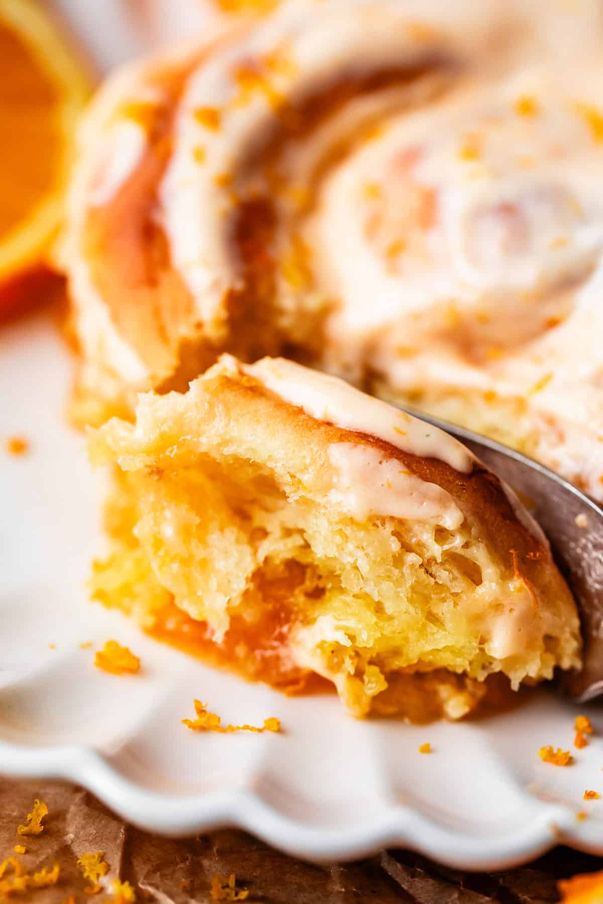A close-up of a partially eaten orange sweet roll with creamy icing and orange zest on a white plate, showing its soft, moist texture and citrus filling.