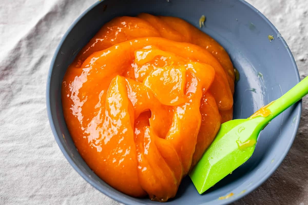 Bright orange, glossy orange roll filling in a gray bowl, with a green silicone spatula resting inside. The bowl is placed on a light-colored, textured fabric surface.
