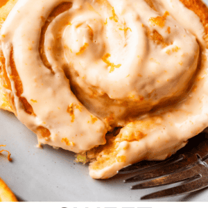 A close-up of a sweet orange roll topped with creamy orange glaze, served on a plate with fresh orange slices in the background. A fork rests nearby. Text at the bottom reads “Sweet Orange Rolls The Food Charlatan.”.