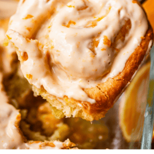 A close-up of a hand lifting a frosted orange roll from a glass baking dish, with fresh orange slices in the background. The orange roll is swirled and topped with creamy orange icing.