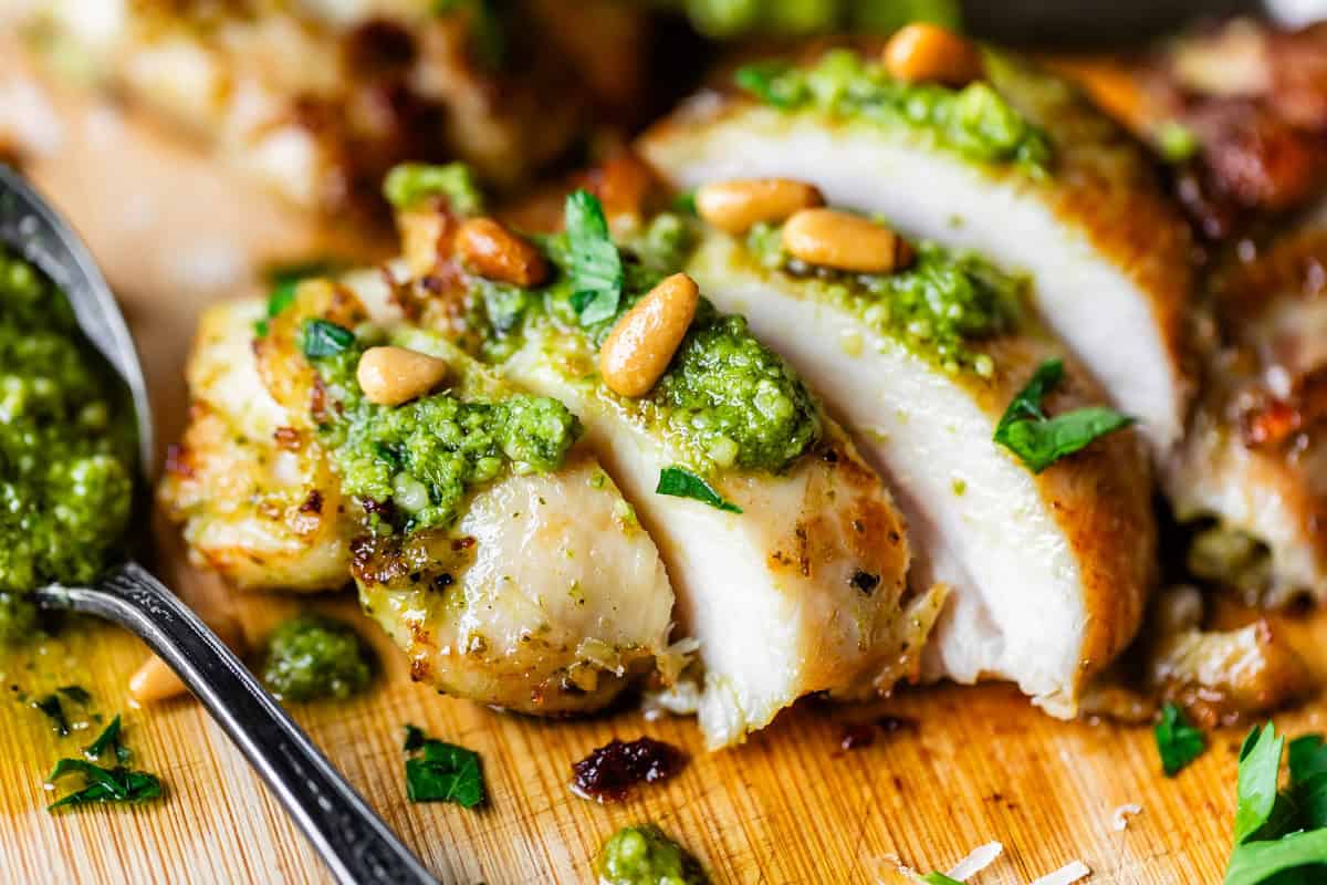 Close-up of sliced grilled chicken breast topped with green pesto sauce, pine nuts, and chopped herbs on a wooden cutting board, with a spoon of pesto beside it.