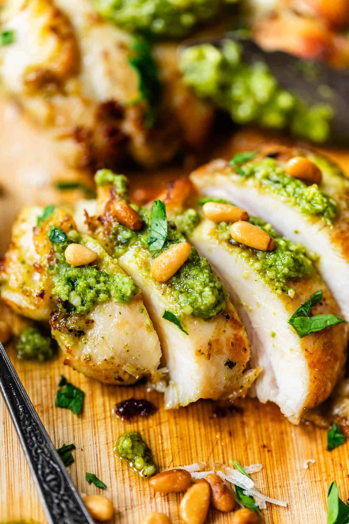 Sliced grilled chicken breast topped with green pesto, pine nuts, and chopped parsley, served on a wooden board.