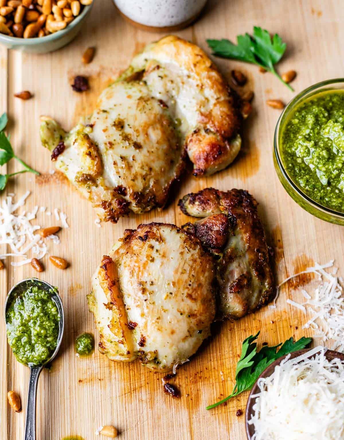 Two grilled chicken thighs sit on a wooden board, topped with green pesto sauce. Grated cheese, pine nuts, parsley, and a bowl of pesto are arranged around the chicken for garnish.