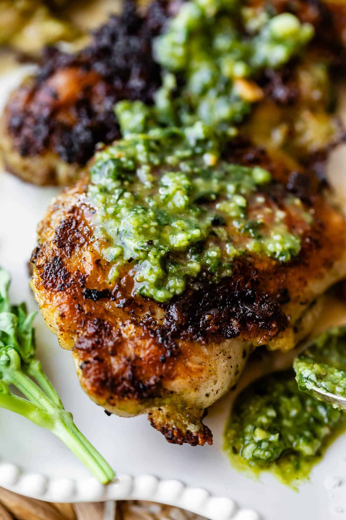Close-up of a piece of grilled chicken topped with green pesto sauce, served on a white plate with a small sprig of fresh herbs on the side. The chicken has a crispy, golden-brown skin.