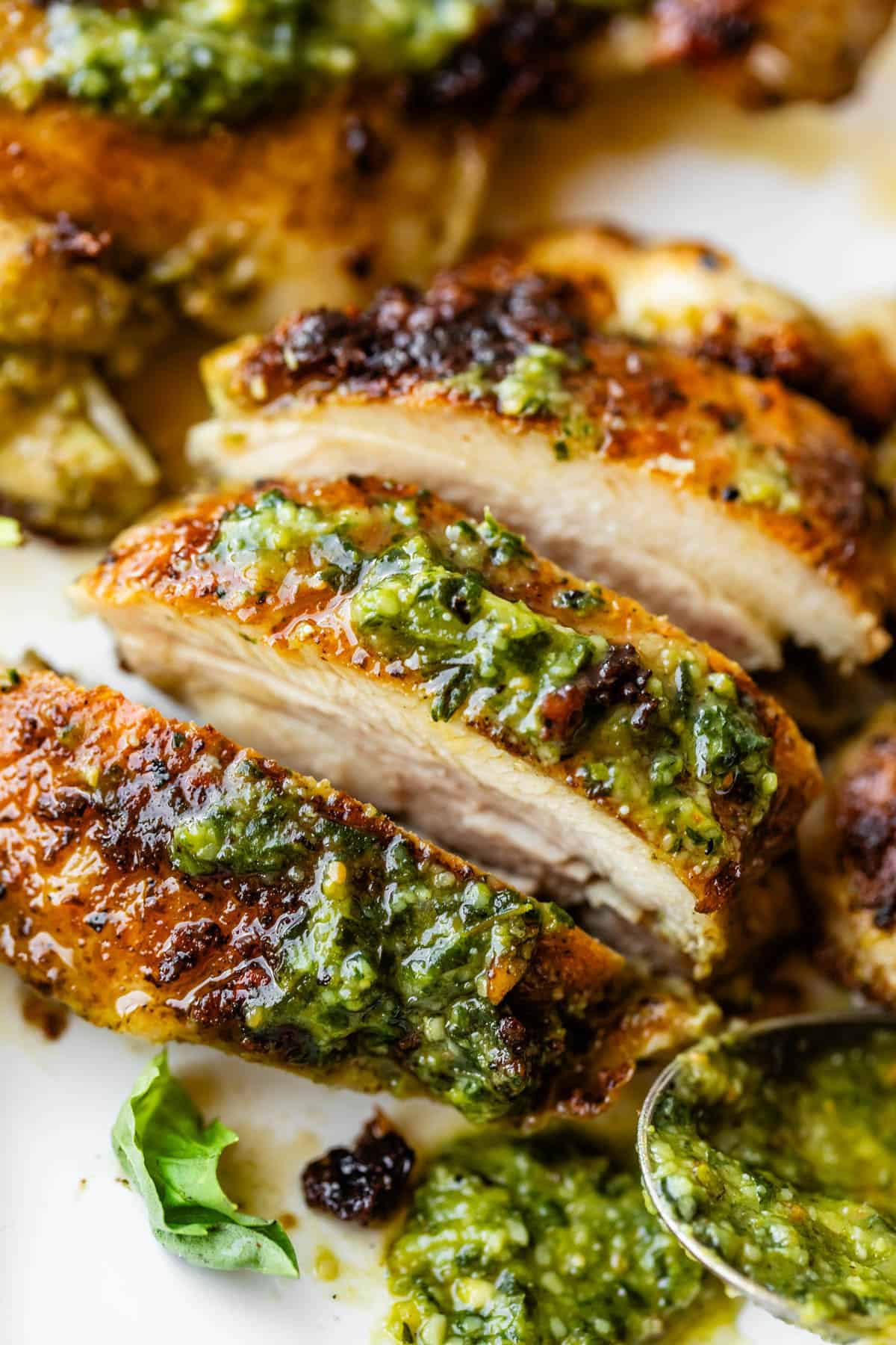 Close-up of sliced, golden-brown grilled chicken breast topped with a vibrant green pesto sauce, garnished with fresh herbs. A spoon with pesto and a basil leaf are visible nearby.