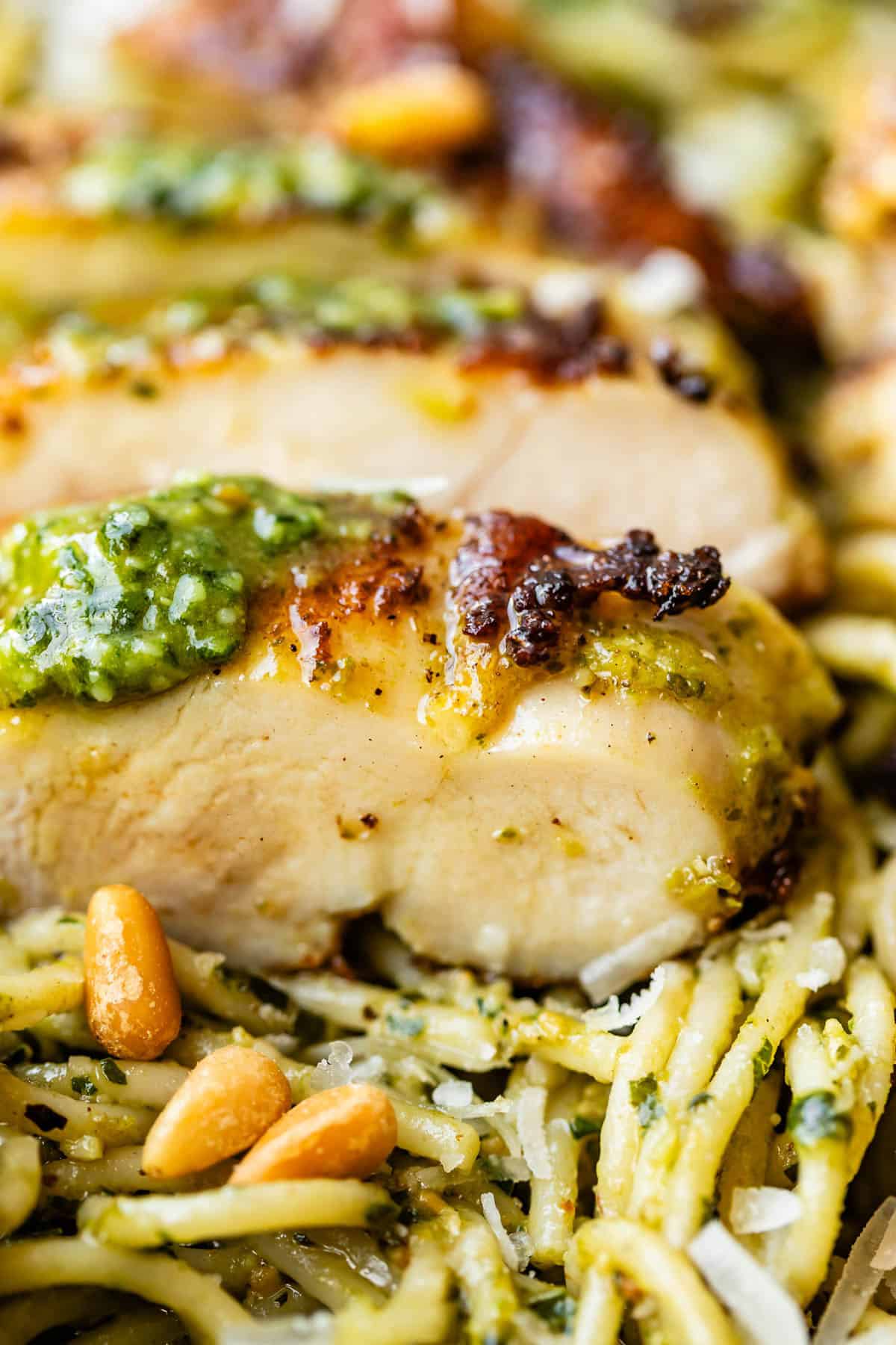 Close-up of sliced, golden-brown chicken breast topped with pesto, resting on a bed of pesto-coated spaghetti with pine nuts and grated cheese.