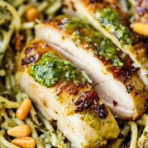 Close-up of sliced grilled chicken breast topped with green pesto sauce, served over spaghetti coated in pesto, and garnished with pine nuts.