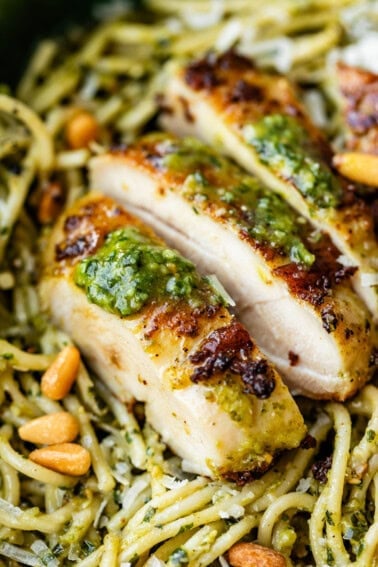 Close-up of sliced grilled chicken breast topped with green pesto sauce, served over spaghetti coated in pesto, and garnished with pine nuts.