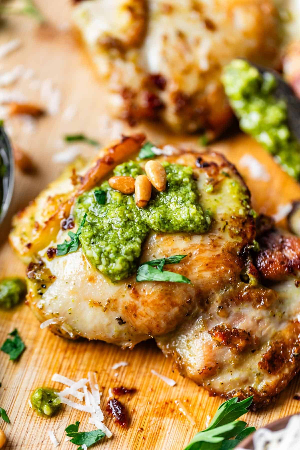 Close-up of a grilled chicken thigh topped with green pesto, pine nuts, and chopped parsley, served on a wooden board with grated cheese and herb garnishes.