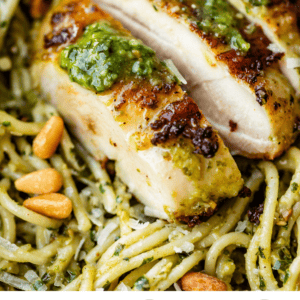 Close-up of sliced, golden-brown chicken breast topped with pesto sauce, served over spaghetti with pesto and pine nuts. Text below reads: Pesto Chicken. Done in 20 minutes! The Food Charlatan.