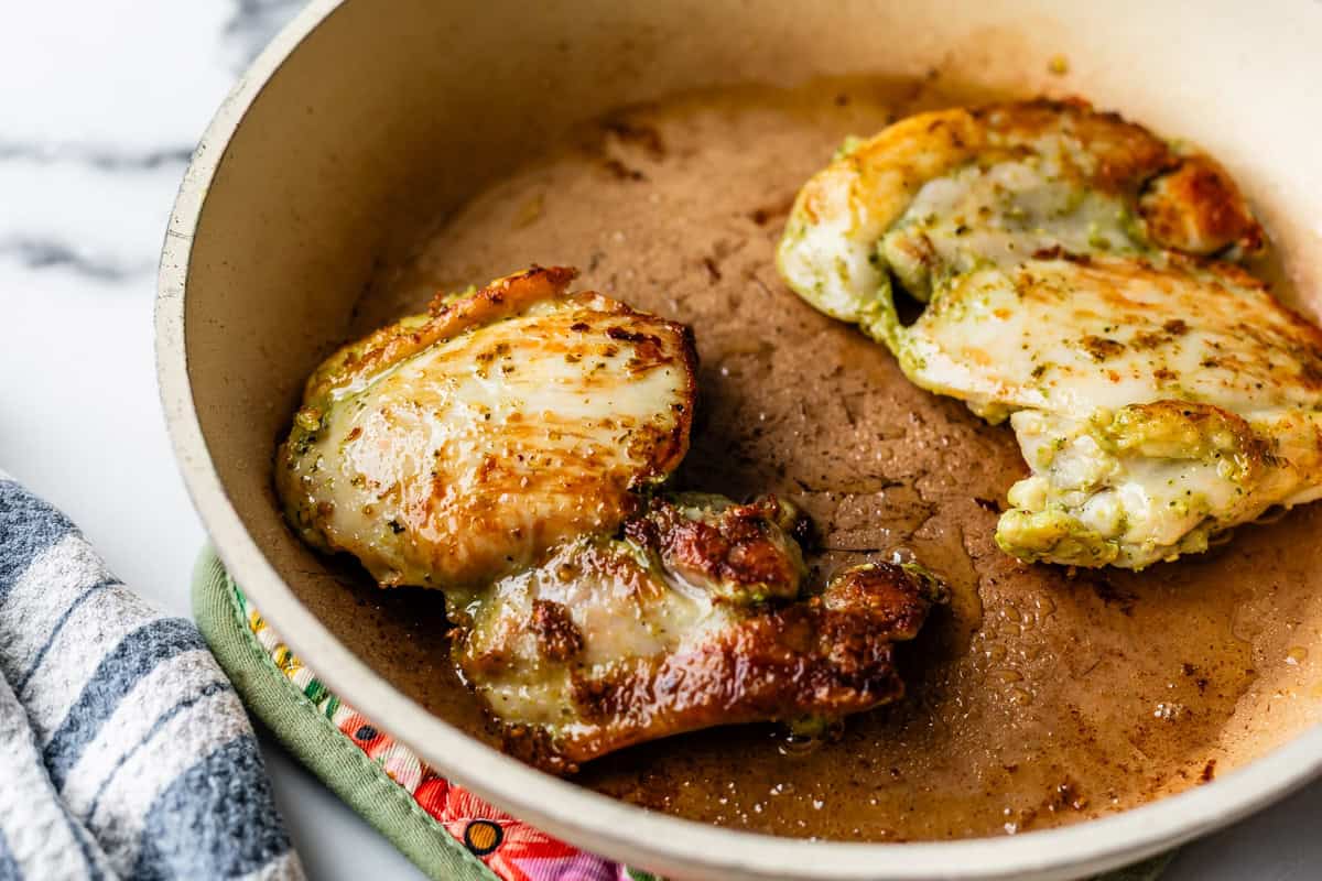 Two browned chicken thighs are cooking in a beige skillet with some oil, resting on a patterned oven mitt. A striped kitchen towel is visible nearby on a marble countertop.